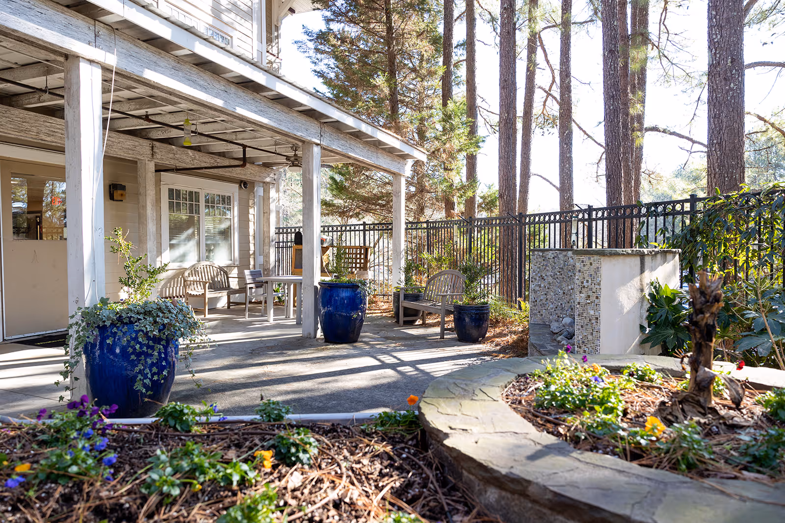 Outdoor patio area at A.G. Rhodes Wesley Woods featuring a covered seating space with wooden benches and chairs, large blue ceramic planters with greenery, a stone-bordered flower bed with colorful flowers, and tall trees in the background behind a black metal fence.