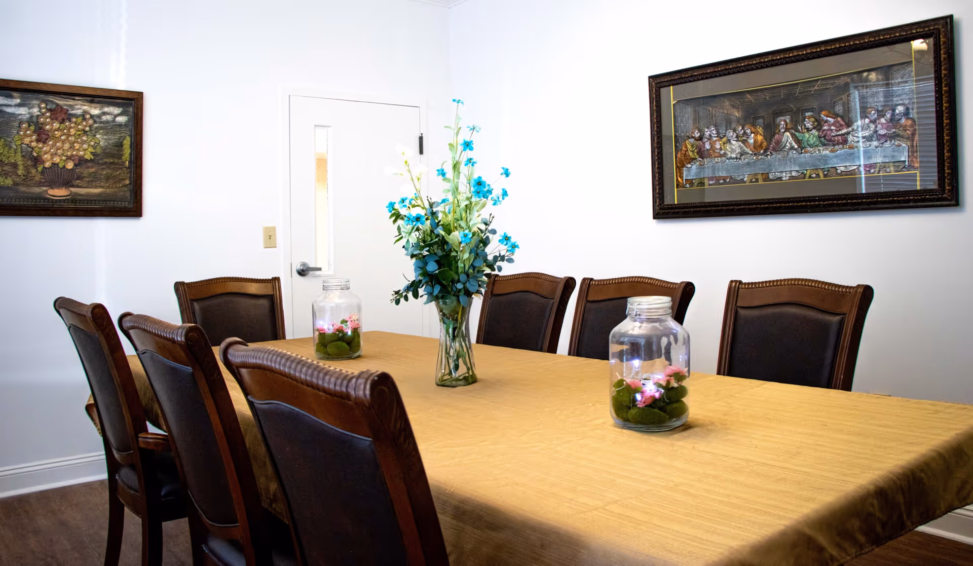 A dining room with a rectangular table covered with a beige tablecloth surrounded by eight wooden chairs with dark cushions. On the table are two glass jars with pink flowers and green moss inside, and a vase with blue and white flowers. The walls are white and decorated with two framed paintings, one depicting grapes and the other a scene resembling The Last Supper.