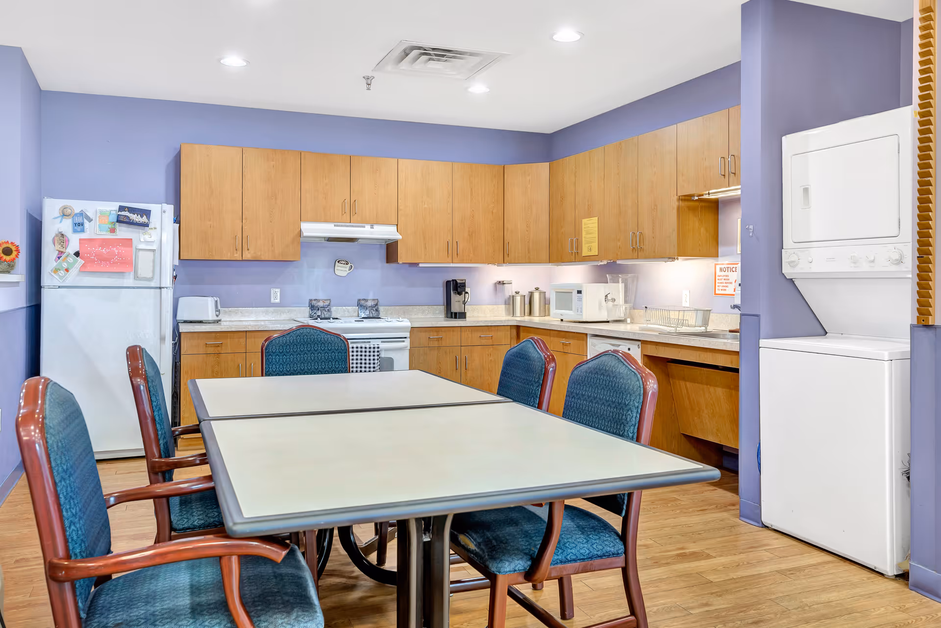 A communal kitchen area with wooden cabinets, a white refrigerator, stove, microwave, coffee maker, and a stacked washer and dryer unit. There is a table with four blue cushioned chairs in the center of the room. The walls are painted light purple and the floor is wooden.