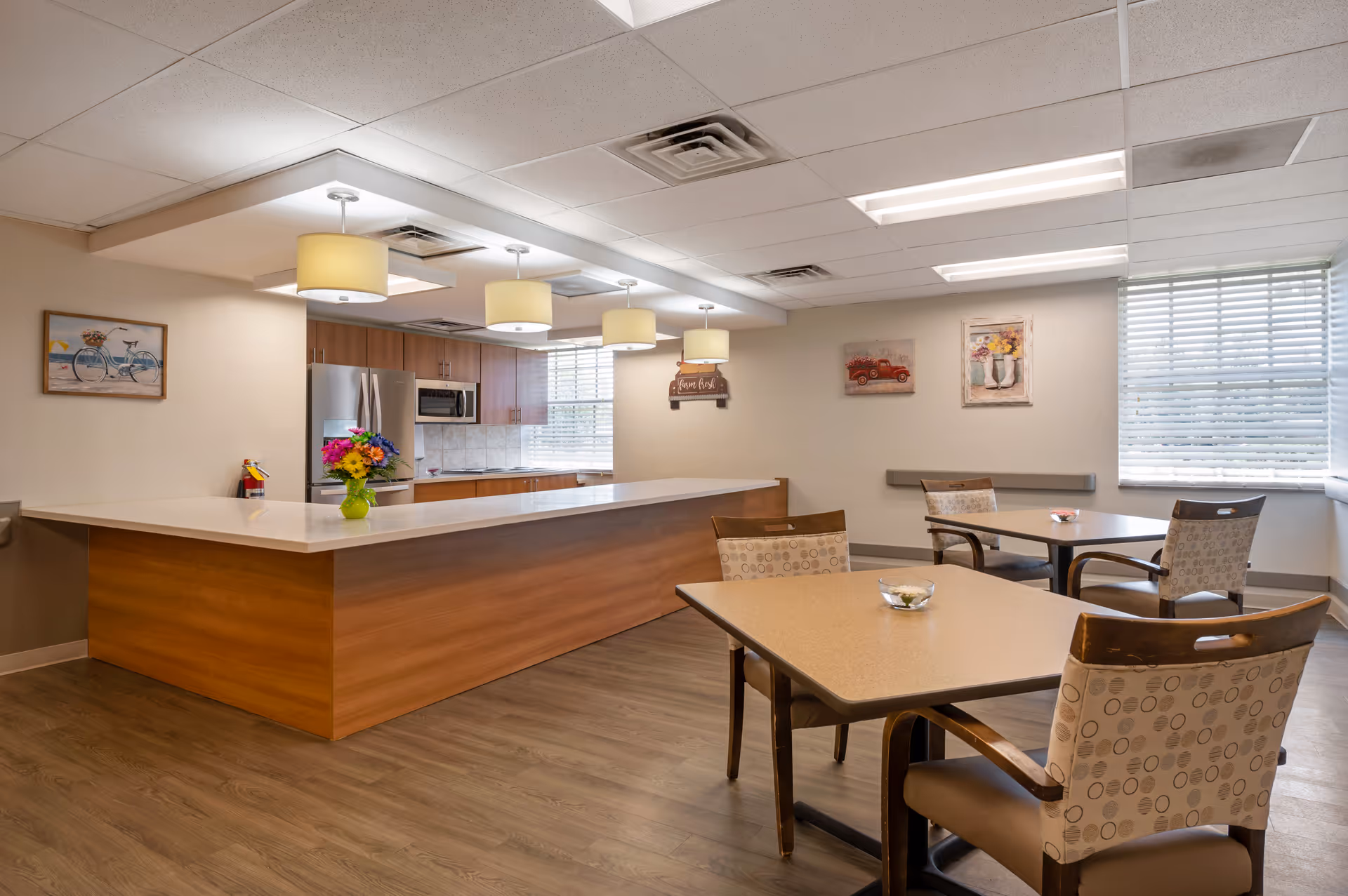A bright and clean dining area in a senior living facility featuring a large wooden counter with a vase of colorful flowers, three pendant lights hanging above, and a kitchen area with stainless steel appliances in the background. There are two tables with four cushioned chairs each, and wall decorations including paintings and a sign. Large windows with blinds allow natural light to fill the room.