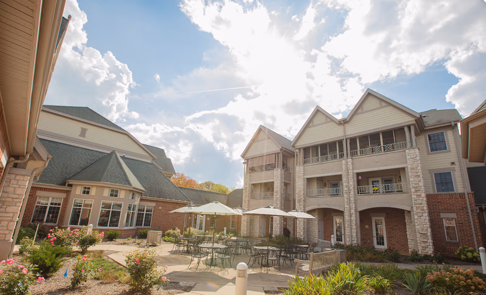 Outdoor courtyard area of a senior living facility with multiple tables and chairs under large umbrellas, surrounded by landscaped gardens and a multi-story building with balconies and large windows under a partly cloudy sky.