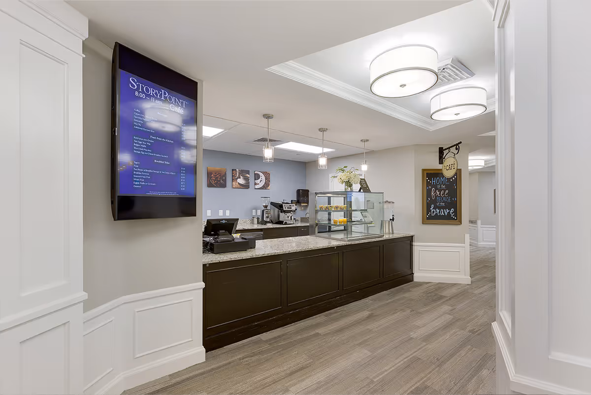 Interior café service counter with a digital menu screen, display case and pendant lights in a senior living community.