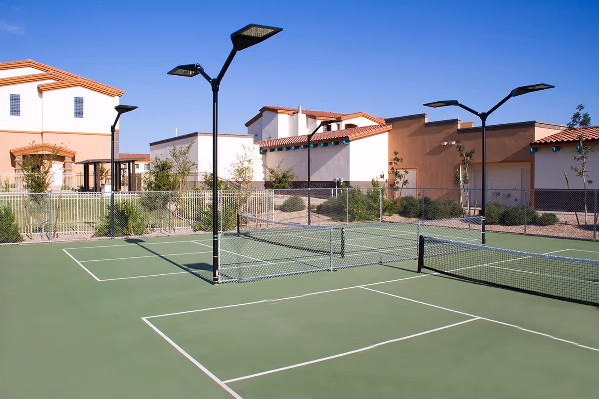 Outdoor tennis or pickleball court with green surface and white boundary lines, surrounded by a chain-link fence. Several tall black light poles are positioned around the court. In the background, there are buildings with beige and brown walls, red tile roofs, and some landscaping with bushes and small trees under a clear blue sky.