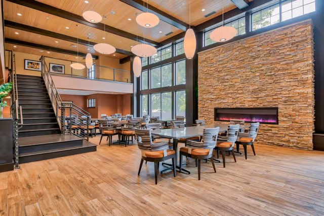 Spacious dining room with multiple tables and chairs, large windows, a stone accent wall with a linear fireplace, and a staircase to a mezzanine.