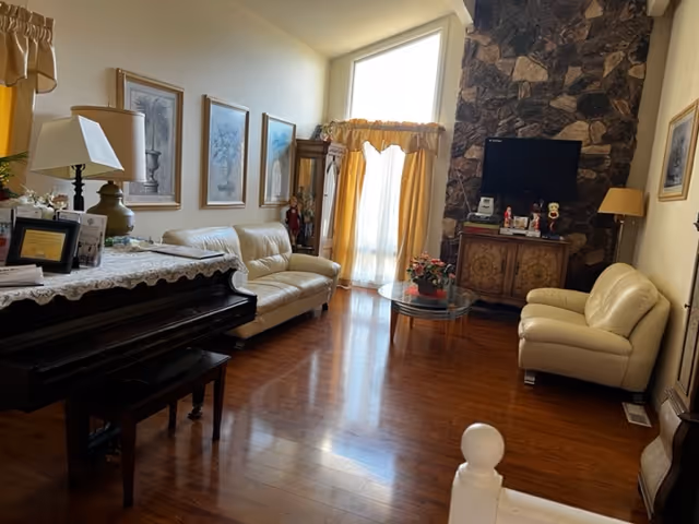 A cozy living room with wooden flooring, two cream-colored leather sofas, a glass coffee table with a flower pot, a stone fireplace with a mounted TV above a wooden cabinet, and a piano with a lace cover and a lamp on it. The room has large windows with yellow curtains allowing natural light to enter.