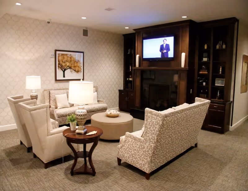 A cozy living room area with patterned wallpaper, two beige armchairs, a patterned loveseat, a round ottoman, and a wooden side table with a lamp and small plant. A dark wooden built-in fireplace with a mounted flat-screen TV above it is visible in the background.
