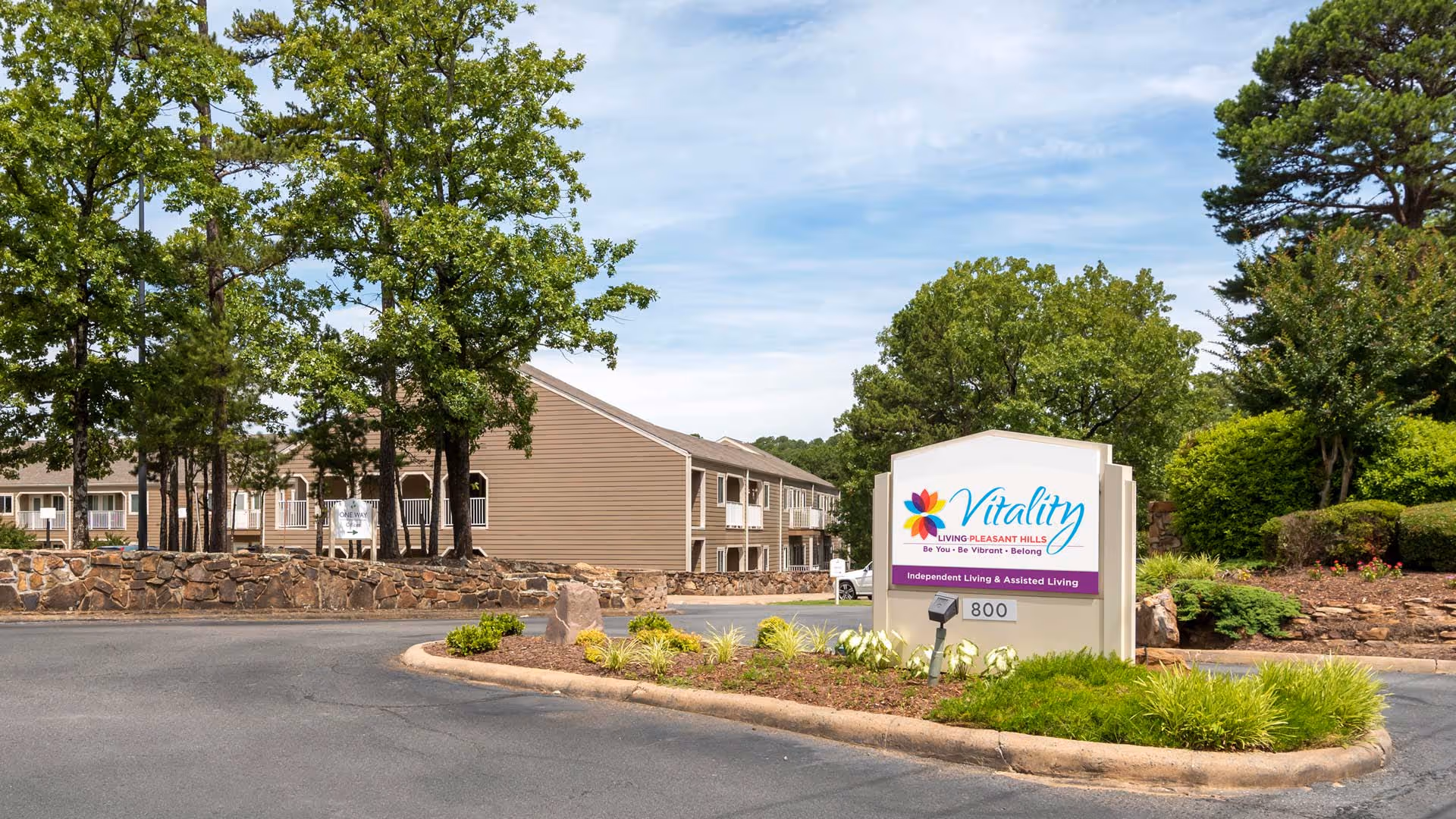Entrance sign for Vitality Living Pleasant Hills, an independent living and assisted living facility, surrounded by landscaped greenery and trees with the facility building visible in the background.