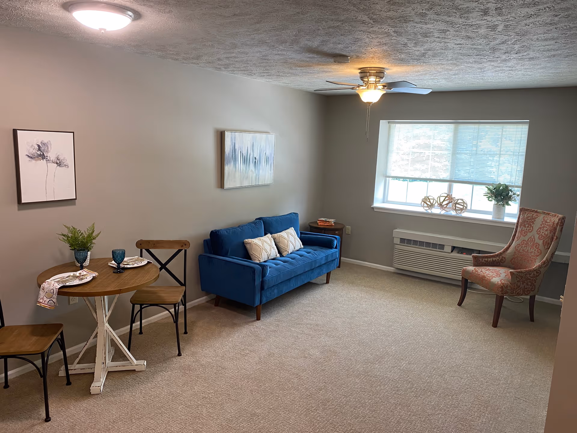 A cozy living room with a blue sofa adorned with two pillows, a patterned armchair near a window with blinds, and a small round wooden dining table with two chairs. The walls are light gray with two pieces of artwork hanging. The room has beige carpet flooring and a ceiling fan with a light.