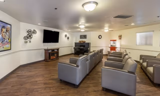 A cozy common room with rows of gray armchairs facing a wall-mounted flat screen TV above a small electric fireplace. The room features wood flooring, a piano in the corner, a popcorn machine, and movie-themed decorations including a Casablanca poster and film reel wall art.