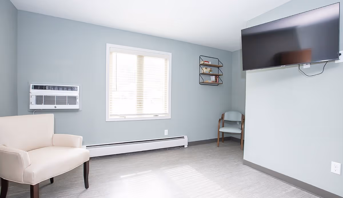 A cozy room featuring a white armchair, a small blue chair, a wall-mounted TV, and a window with blinds, all set against a light blue wall.