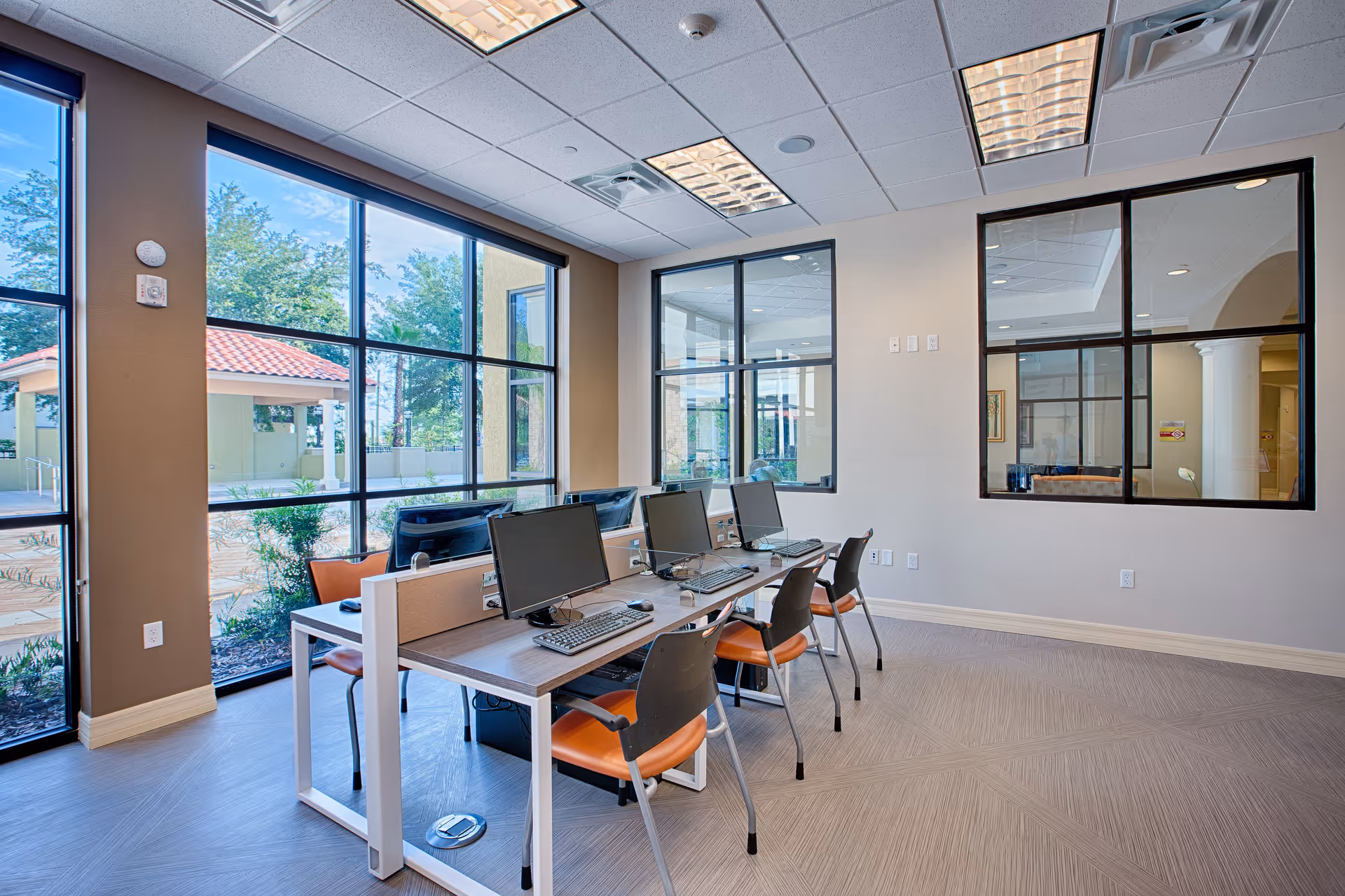 A bright computer room with large windows showing an outdoor view. The room has a long table with four desktop computers and chairs with orange seats. The walls have large interior windows looking into another room.