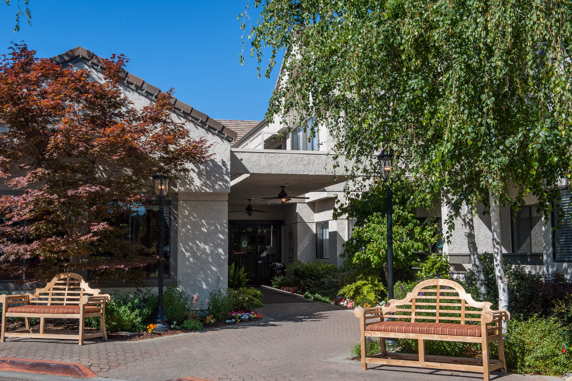 Entrance of a senior living facility with a covered drop-off, two wooden benches, and trees and landscaping.
