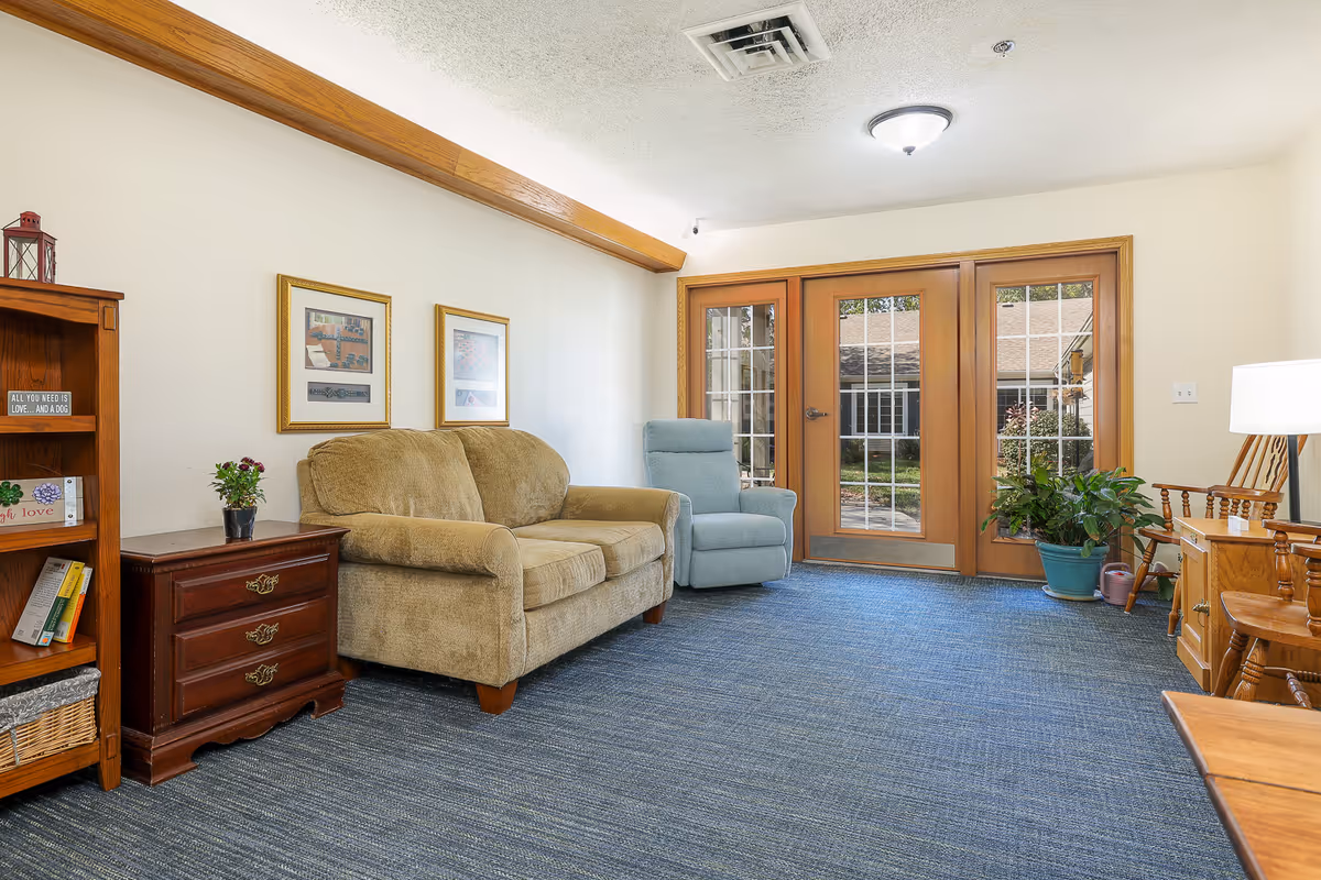 Comfortable seating area with a beige sofa, blue recliner, wooden furniture, plants, and glass patio doors to an outdoor courtyard.