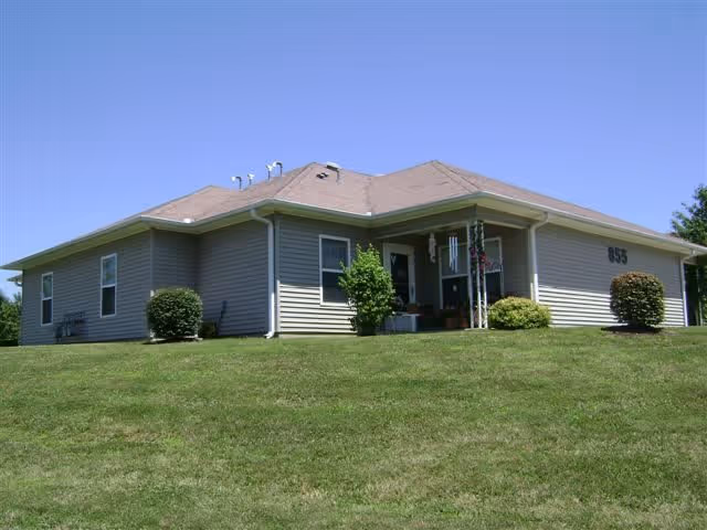 Single-story residential building with beige siding and a light brown roof, surrounded by a well-maintained grassy lawn and a few bushes, under a clear blue sky.