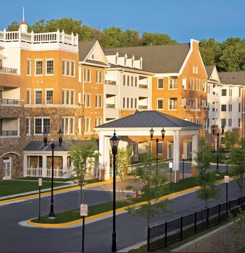Exterior view of a multi-story brick and stone retirement community building with a covered entrance driveway, street lamps, small trees, and a clear blue sky in the background.