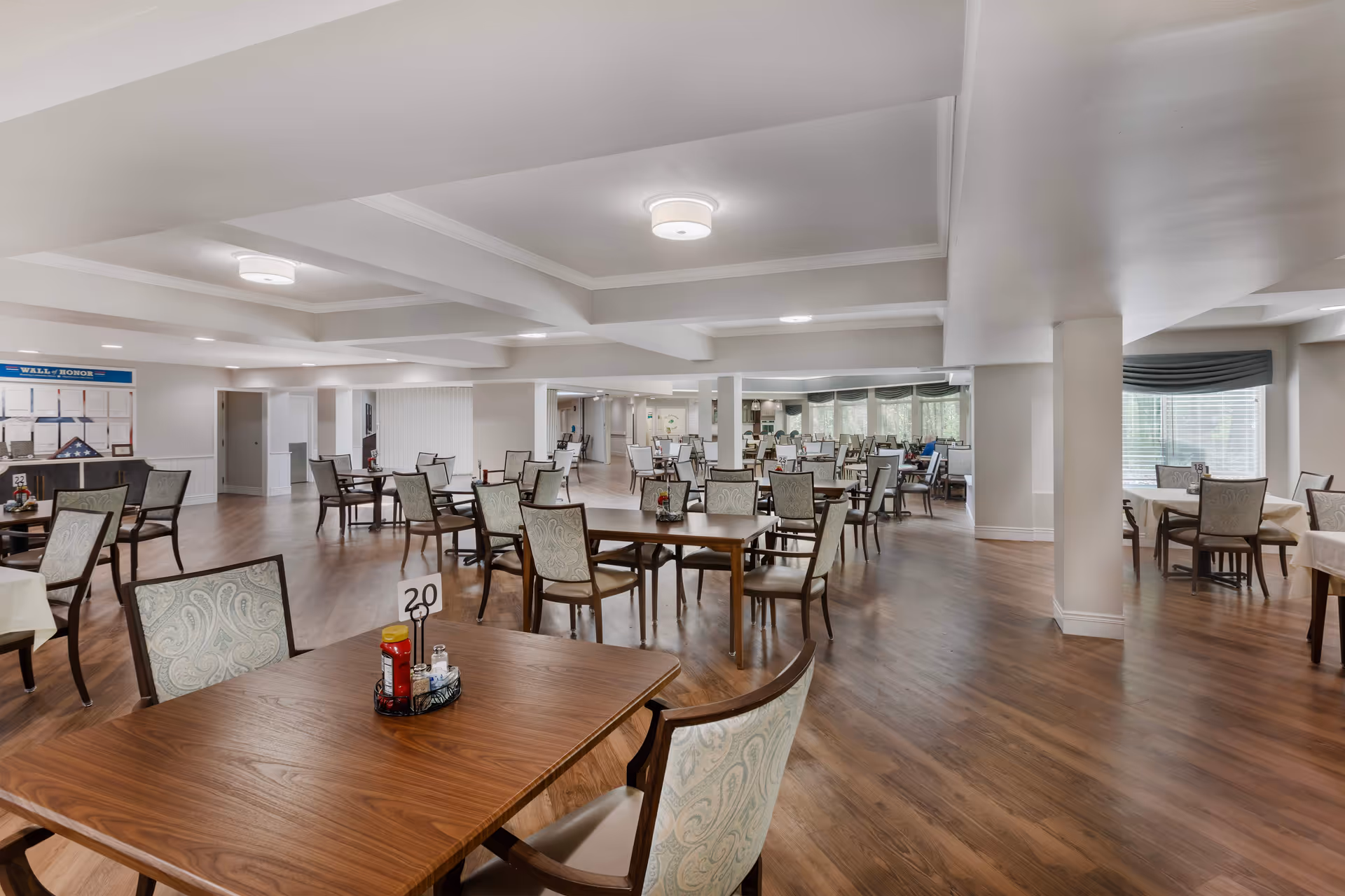 Spacious senior living dining room with multiple wooden tables and upholstered chairs on hardwood floors.