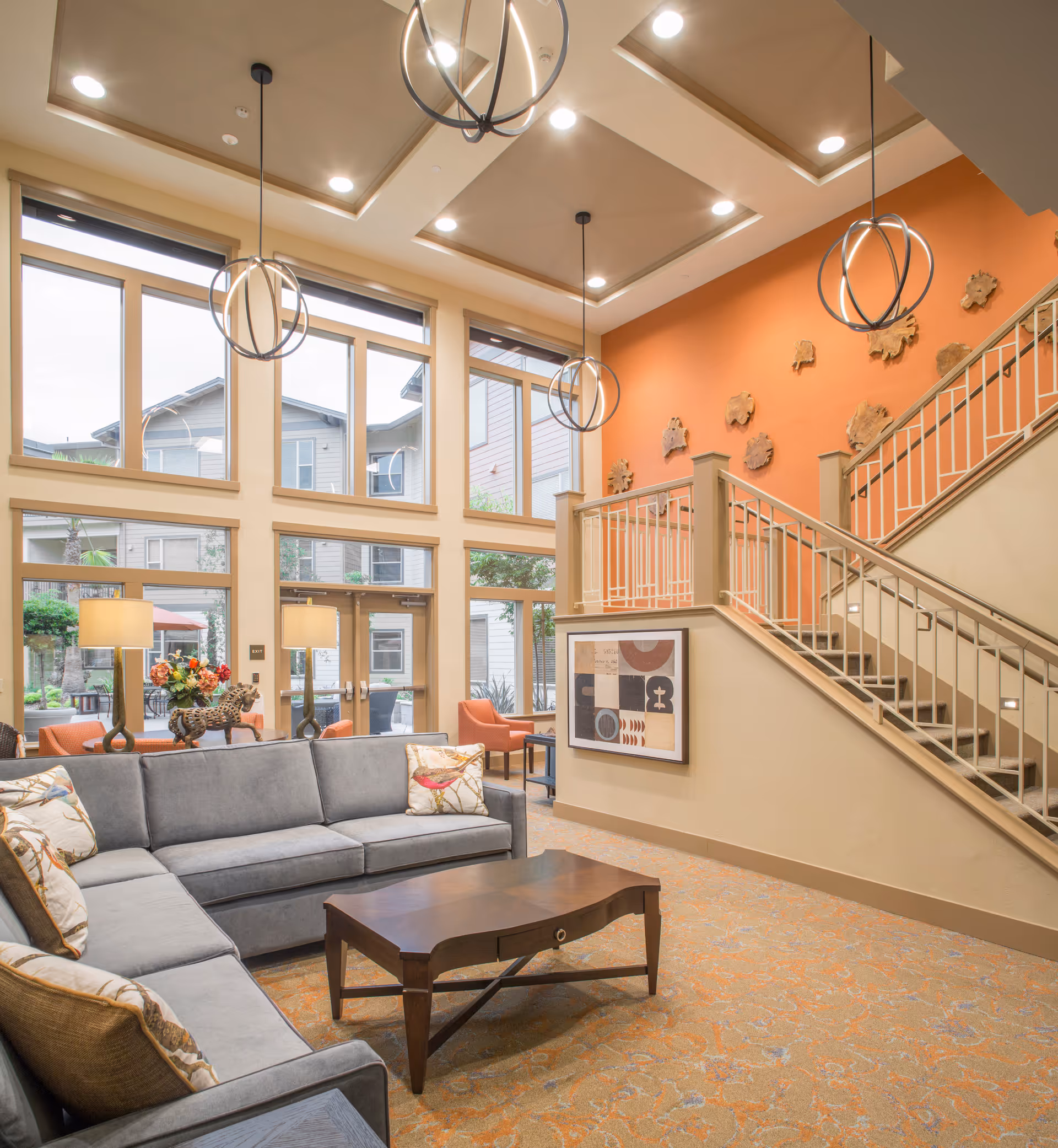 A bright and spacious living room area with a large gray sectional sofa adorned with decorative pillows, a wooden coffee table, and modern pendant lights hanging from a high ceiling. The room features large windows letting in natural light, an orange accent wall with decorative art pieces, and a staircase with a beige railing. Outside the windows, parts of the building and greenery are visible.
