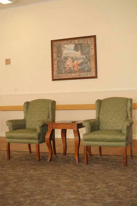 Two green upholstered armchairs flank a wooden side table under a framed painting.