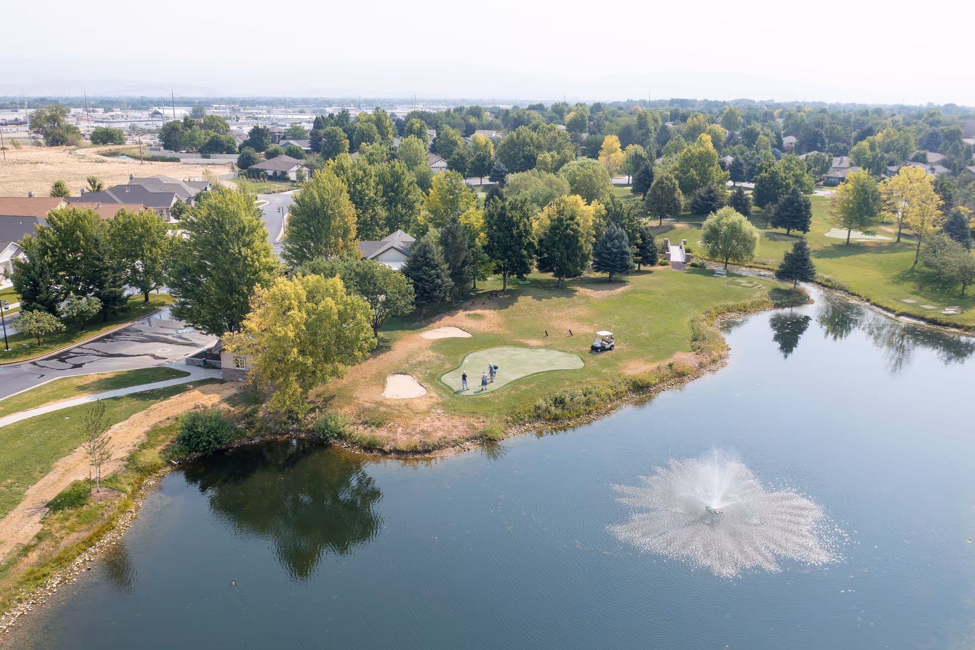 Aerial view of a green park area with a small golf putting green, surrounded by trees and a pond with a water fountain. There are a few people near the putting green and a golf cart nearby. Residential houses and roads are visible in the background under a clear sky.