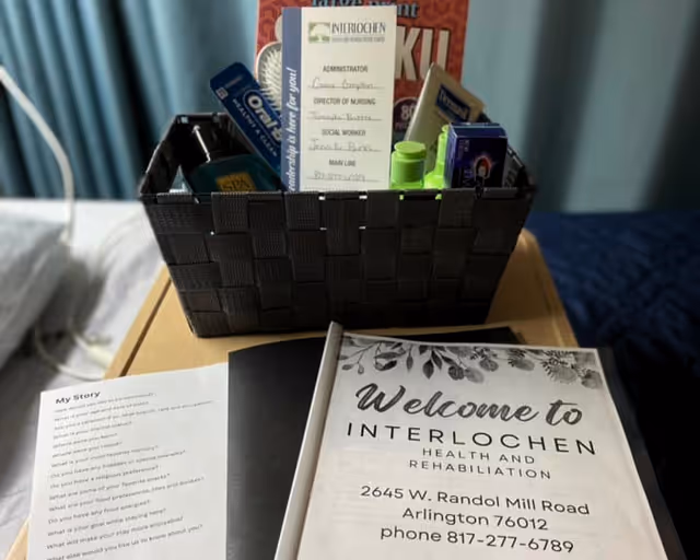 A black woven basket on a bedside table containing various personal care items and a card listing staff names at Interlochen Health and Rehabilitation Center. In front of the basket is a welcome booklet with the facility's name, address, and phone number. Part of a bed and a blue curtain are visible in the background.