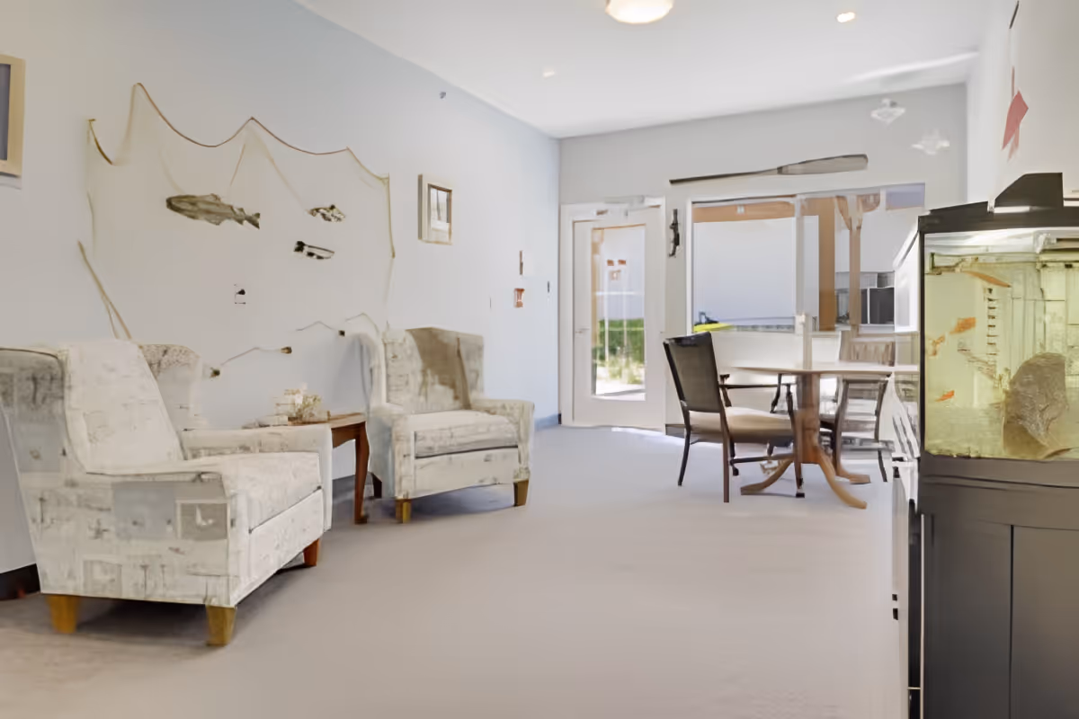 Bright common room with upholstered armchairs, a round dining table and chairs, and a fish tank near a glass door.