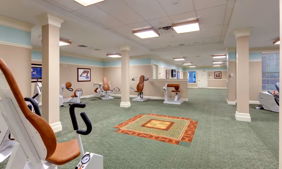 A spacious exercise room with green carpet and beige walls featuring several pieces of fitness equipment including stationary bikes and strength training machines. The room has white ceiling tiles with recessed lighting and decorative columns. There are framed pictures on the walls and a window with blinds on the right side.