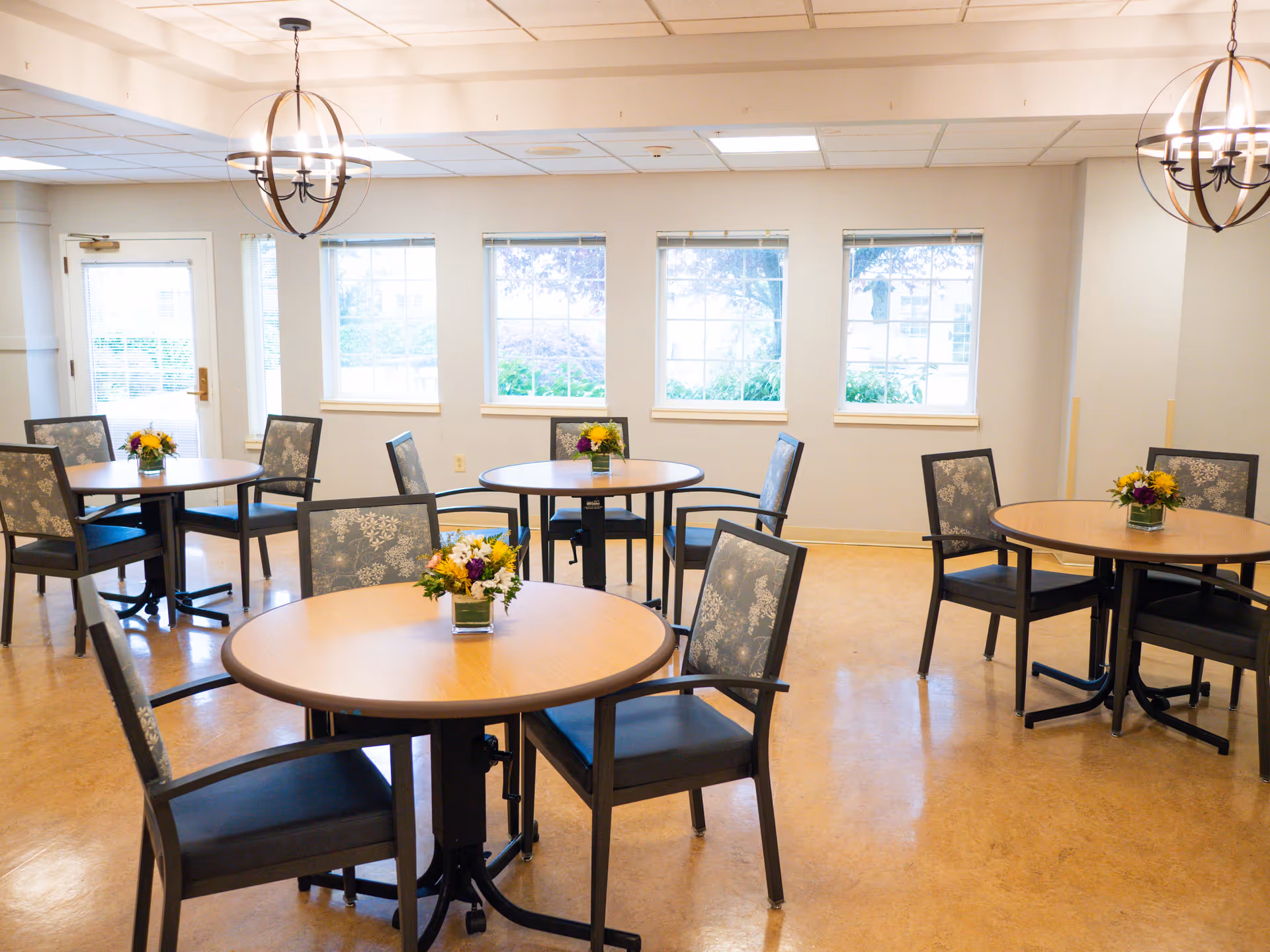 A bright dining room with round wooden tables and chairs arranged neatly. Each table has a small floral centerpiece. The room has large windows letting in natural light and two modern chandeliers hanging from the ceiling.