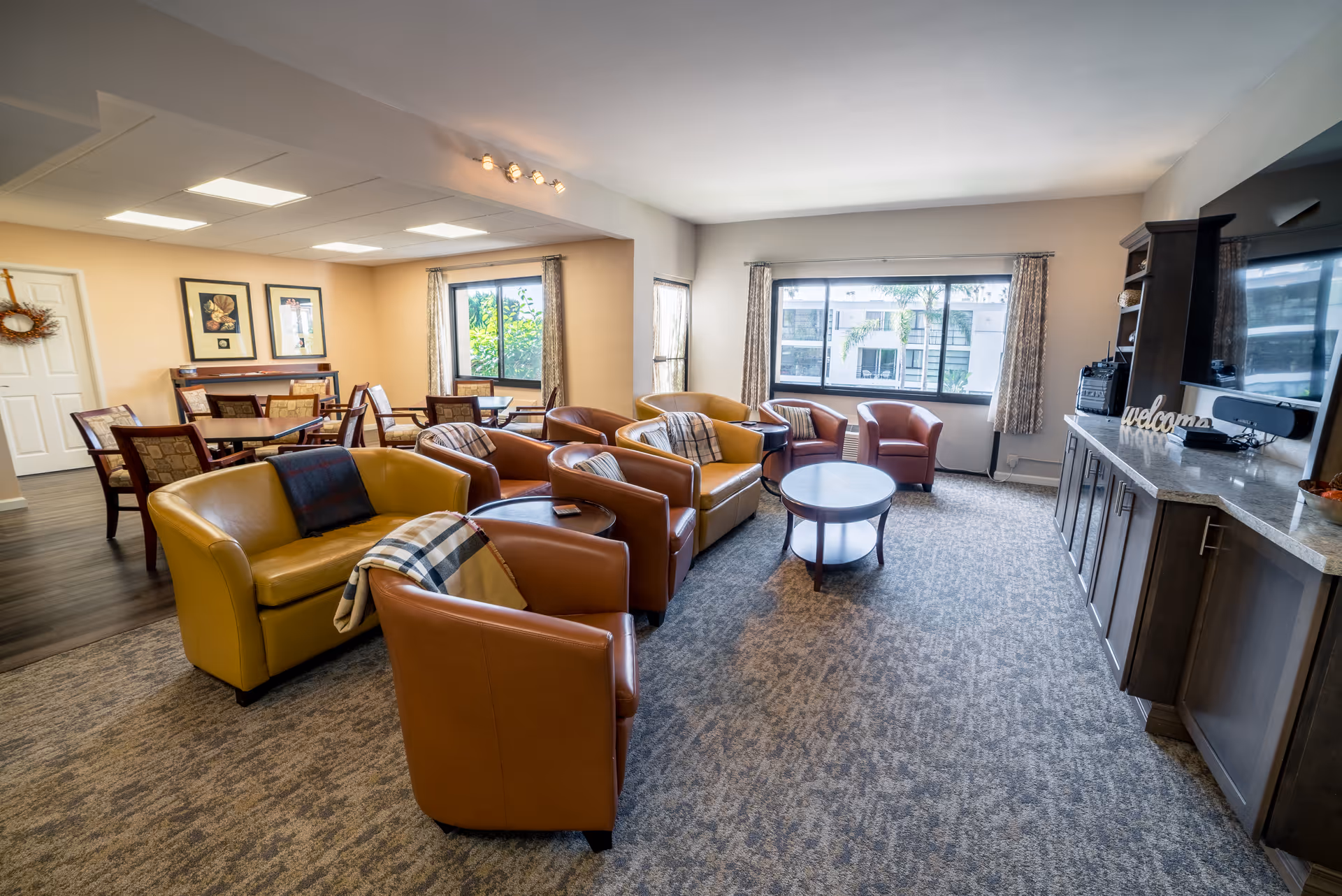 A bright and spacious living room area with multiple leather armchairs and sofas arranged around a round wooden coffee table. Large windows with curtains let in natural light, and a cabinet with a television and decorative items is against one wall. In the background, there is a dining area with tables and chairs, and framed artwork on the walls.