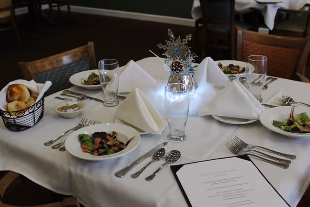A round dining table set with white tablecloth, folded white napkins, glasses, silverware, salad plates with greens, a basket of bread rolls, and a festive centerpiece with pine cones and silver decorations.