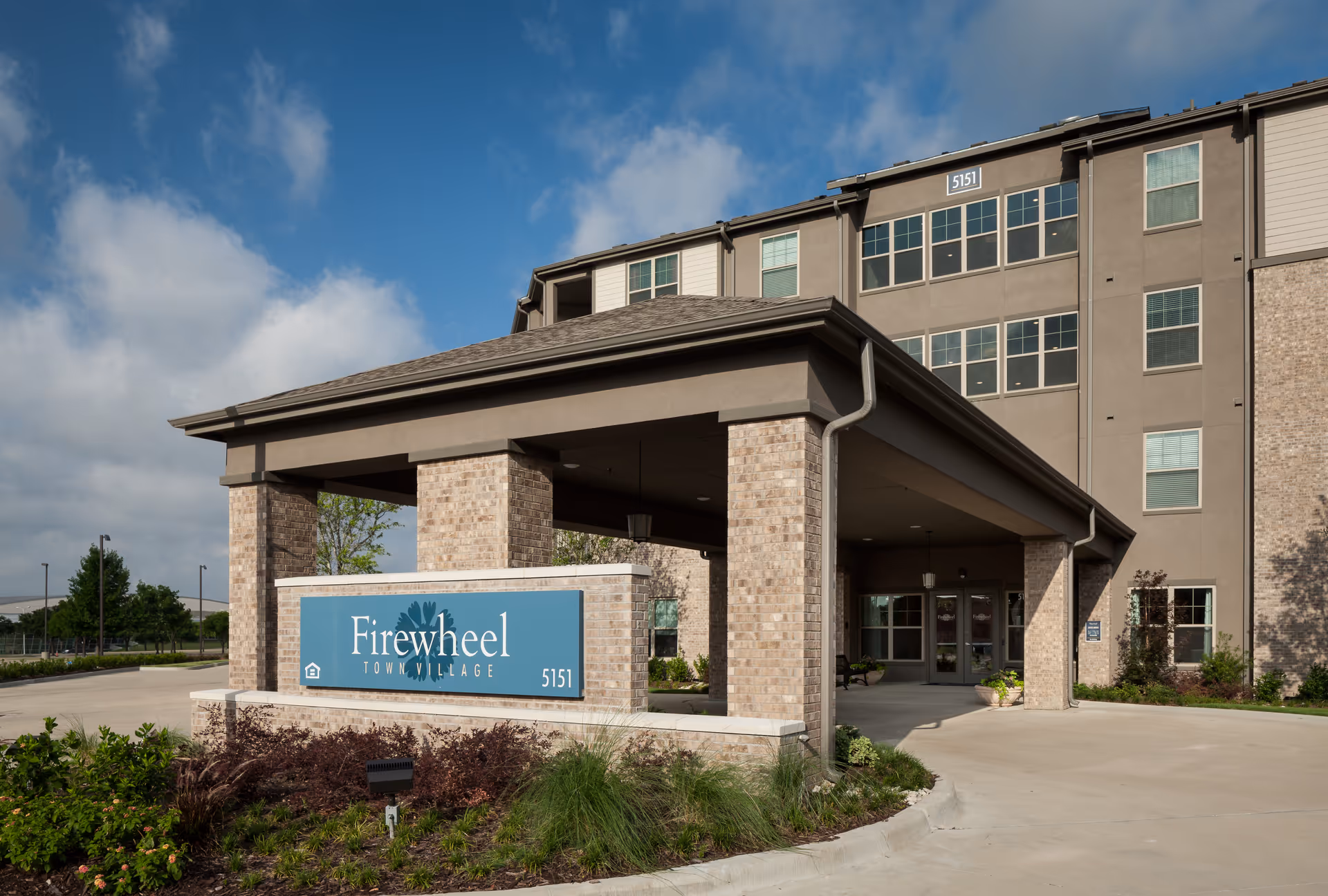 Entrance of Firewheel Town Village senior living facility showing a covered drop-off area with brick pillars, a blue sign with the facility name and address number 5151, and a multi-story building in the background under a partly cloudy sky.