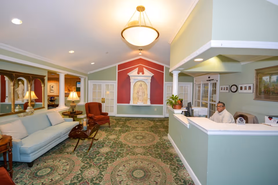 Interior view of a senior living facility lobby with a green patterned carpet, light green walls, a blue sofa, a red armchair, a wooden coffee table, a reception desk with a staff member seated behind it, and a decorative statue set in a red alcove on the far wall.
