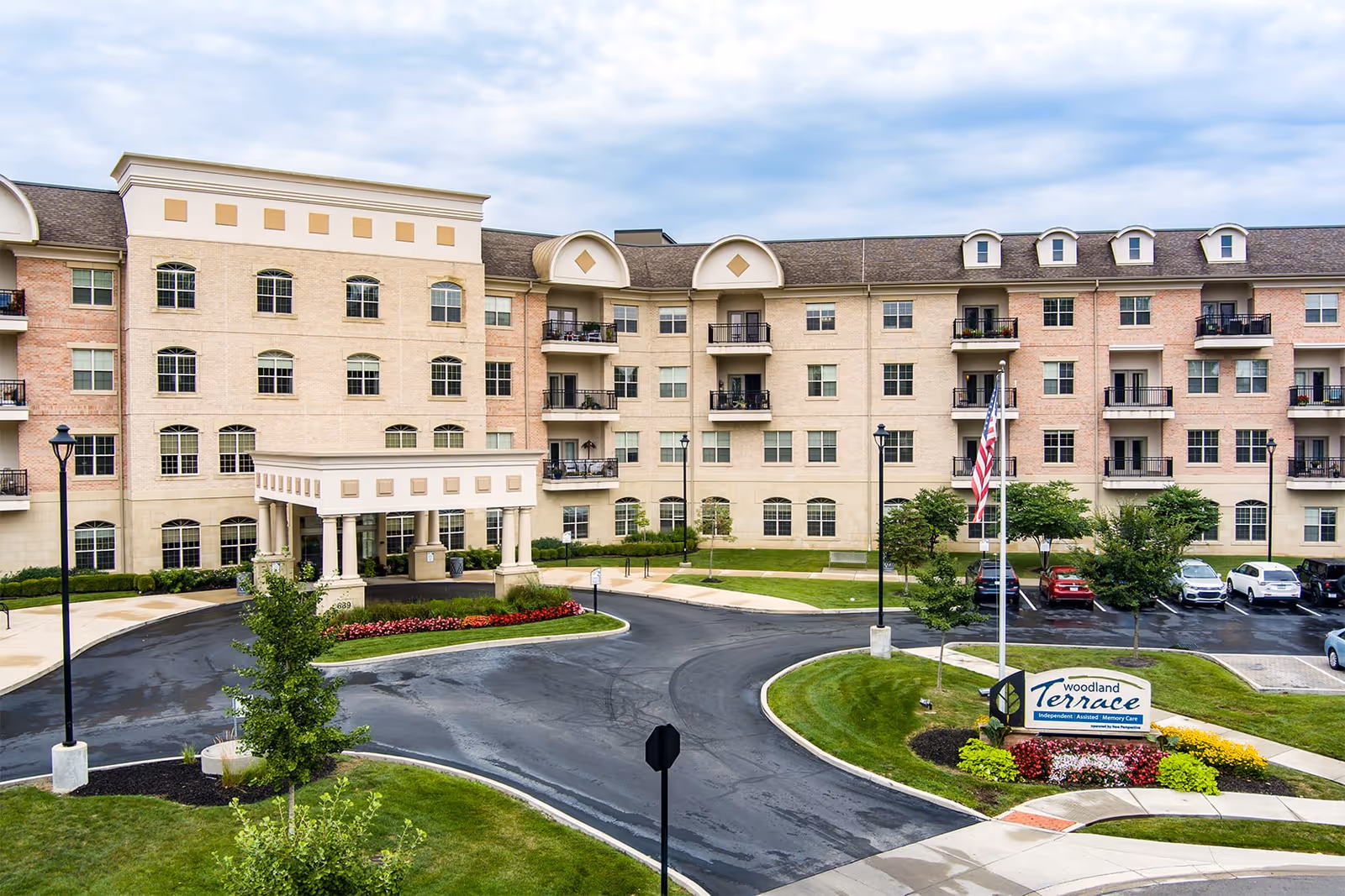 Exterior view of a multi-story senior living facility named Woodland Terrace of Carmel, featuring a large entrance with columns, multiple balconies, a circular driveway, landscaped greenery, and a parking area with several cars.