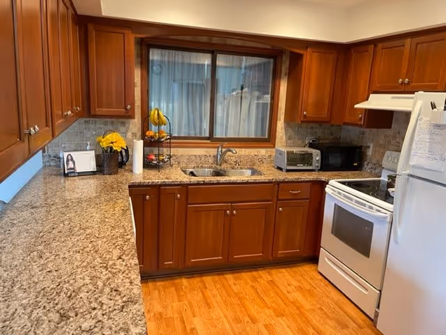 A kitchen with wooden cabinets, granite countertops, a double sink under a window, a white stove, a white refrigerator, a microwave, a toaster oven, a paper towel holder, a fruit stand with bananas, and a vase with yellow flowers on the counter.