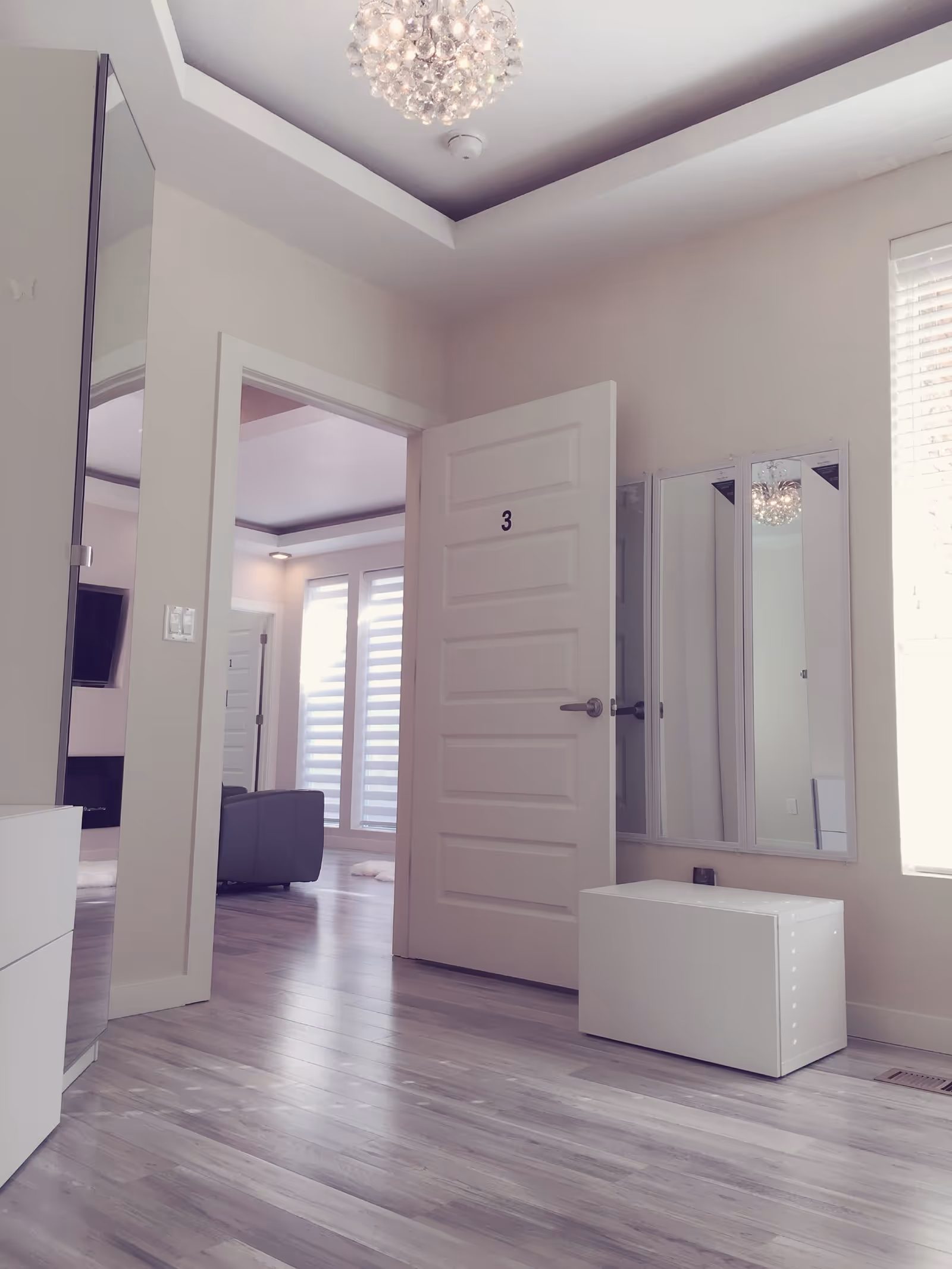 Interior view of a modern room with light-colored walls and wood flooring. A white door labeled with the number 3 is open, revealing a glimpse of a living area with a gray chair and large windows with blinds. The room features a decorative ceiling light fixture, a white storage bench, and three vertical mirrors mounted on the wall next to the door.
