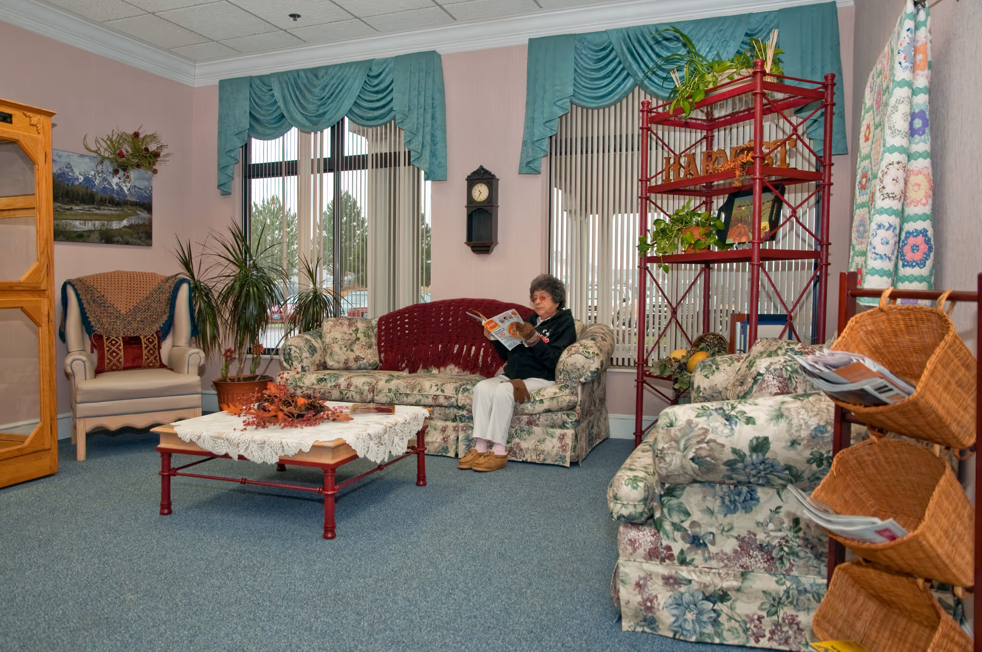 An elderly woman sits on a floral sofa reading a magazine in a cozy living room with armchairs, a coffee table, plants, and shelving.