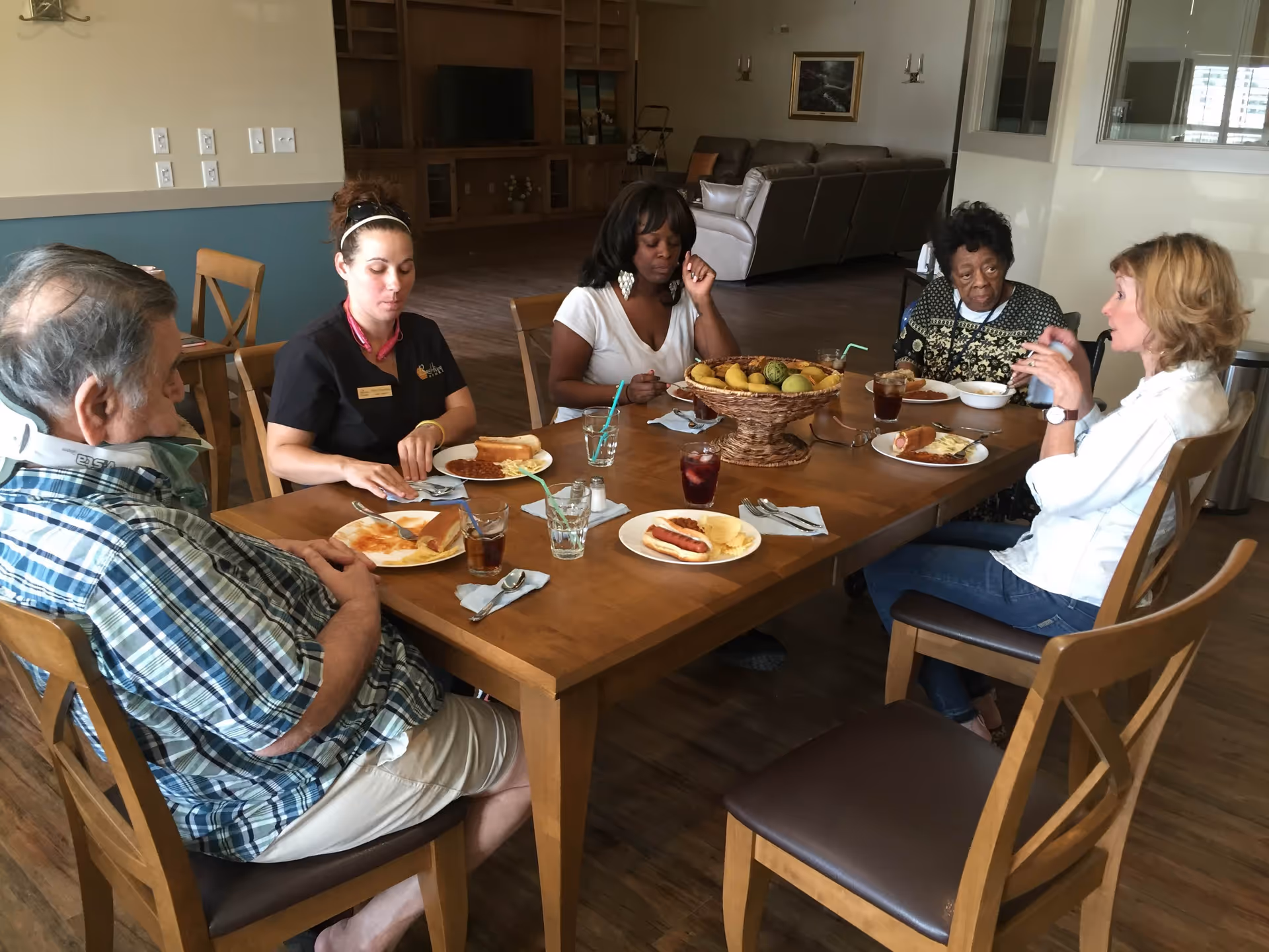Five people sitting around a wooden dining table in a senior living facility, eating a meal that includes hot dogs, chips, and drinks. The room has wooden flooring, a television in the background, and comfortable seating areas.