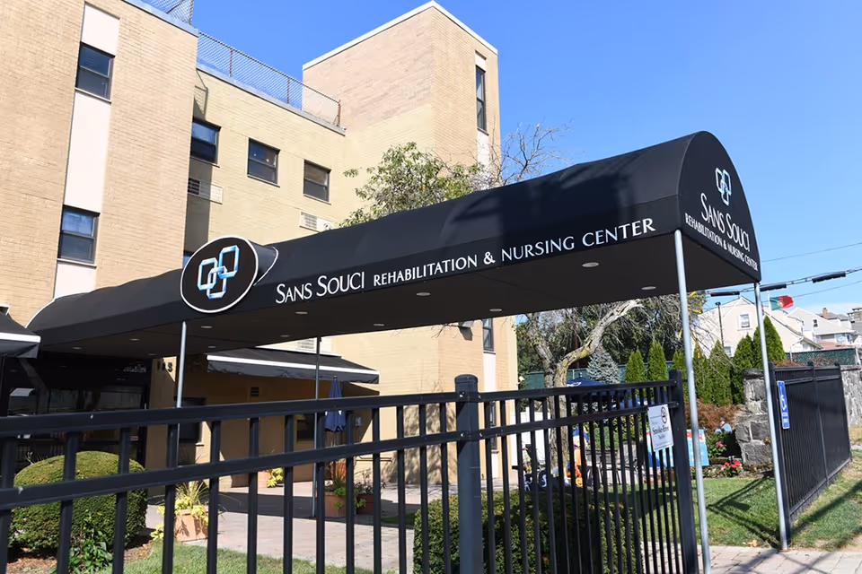 Entrance canopy of Sans Souci Rehabilitation and Nursing Center with the facility's name and logo displayed on a black awning. The building is a multi-story beige brick structure with several windows. There is a black metal fence in the foreground and some greenery around the entrance area under a clear blue sky.
