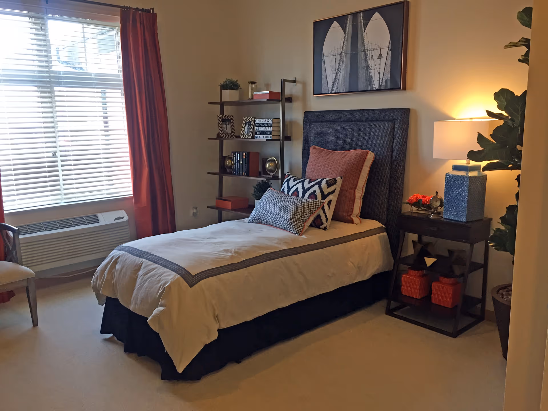 A neatly made single bed with multiple decorative pillows in a cozy bedroom. Next to the bed is a wooden nightstand with a blue lamp, a small clock, and decorative items. A tall shelf with books and framed pictures stands against the wall. A large window with white blinds and red curtains lets in natural light. The room has beige carpet and a framed black and white picture above the bed.