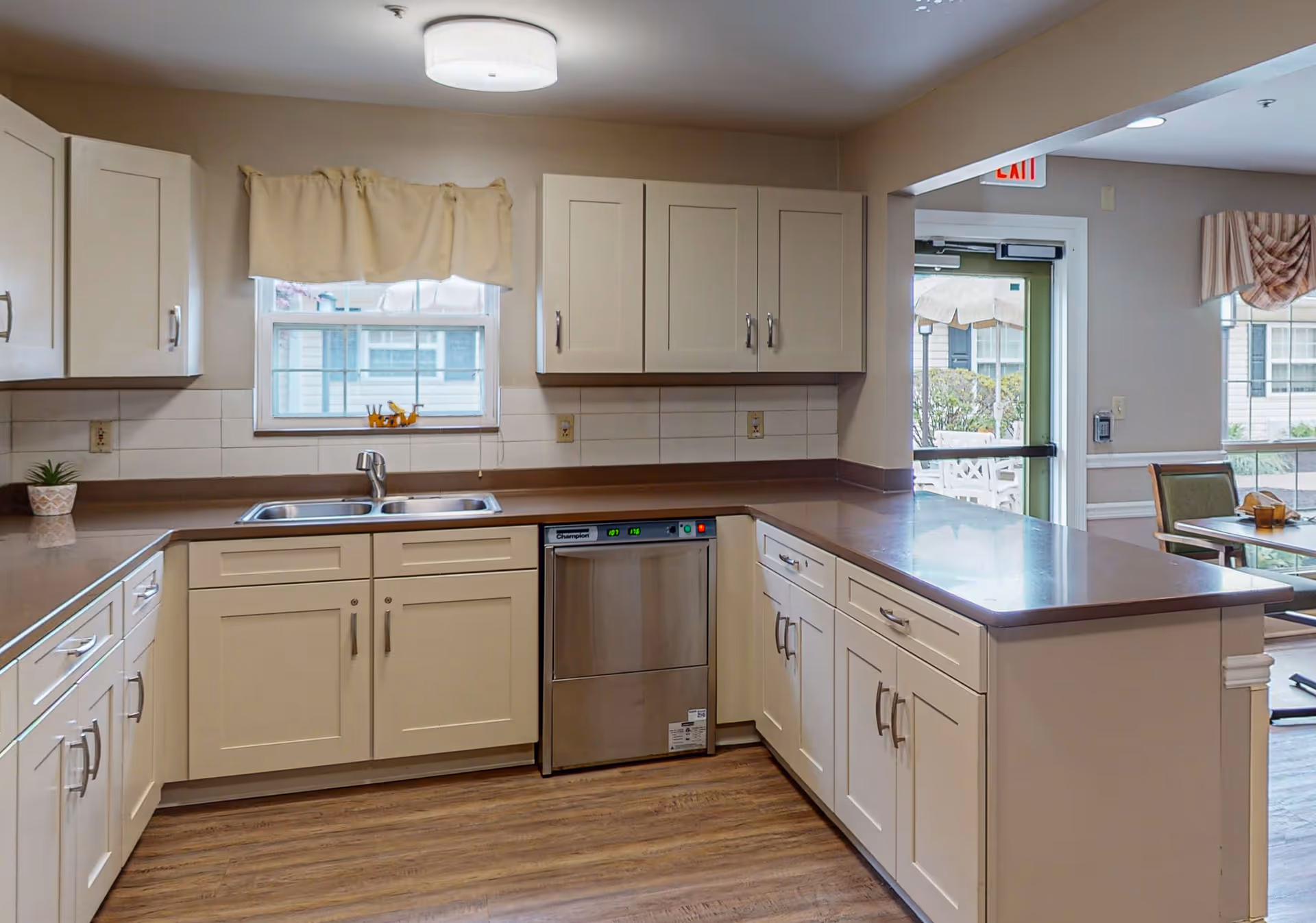 Bright communal kitchen with cream cabinets, stainless steel sink and dishwasher and a large counter opening to a dining area.