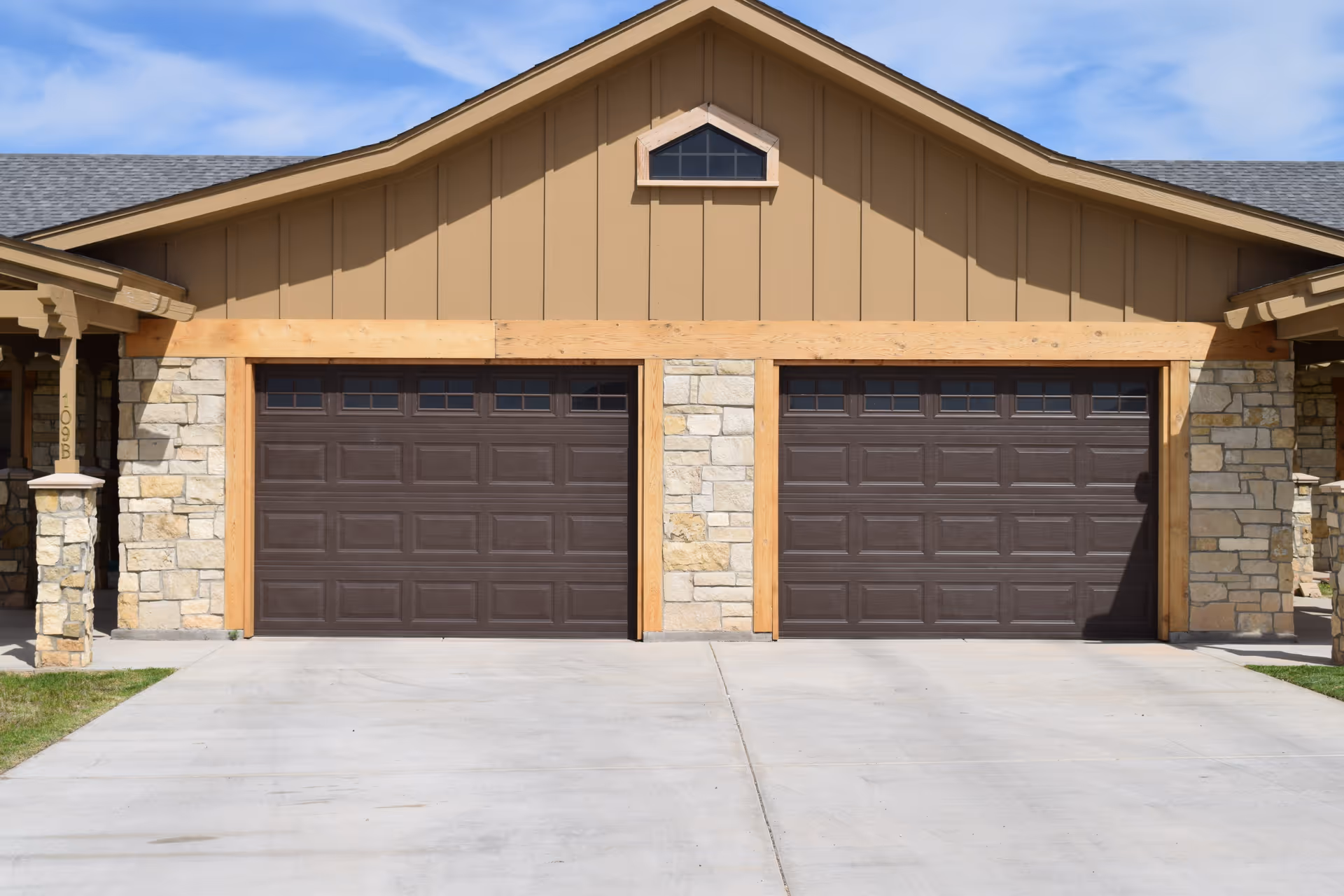 Two closed dark brown garage doors in a stone-and-wood building facade with a concrete driveway.