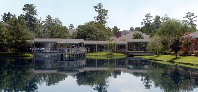 A serene outdoor scene featuring a calm pond with clear reflections of surrounding trees and buildings. There is a covered wooden gazebo extending over the water and a bridge connecting to a building complex surrounded by greenery.