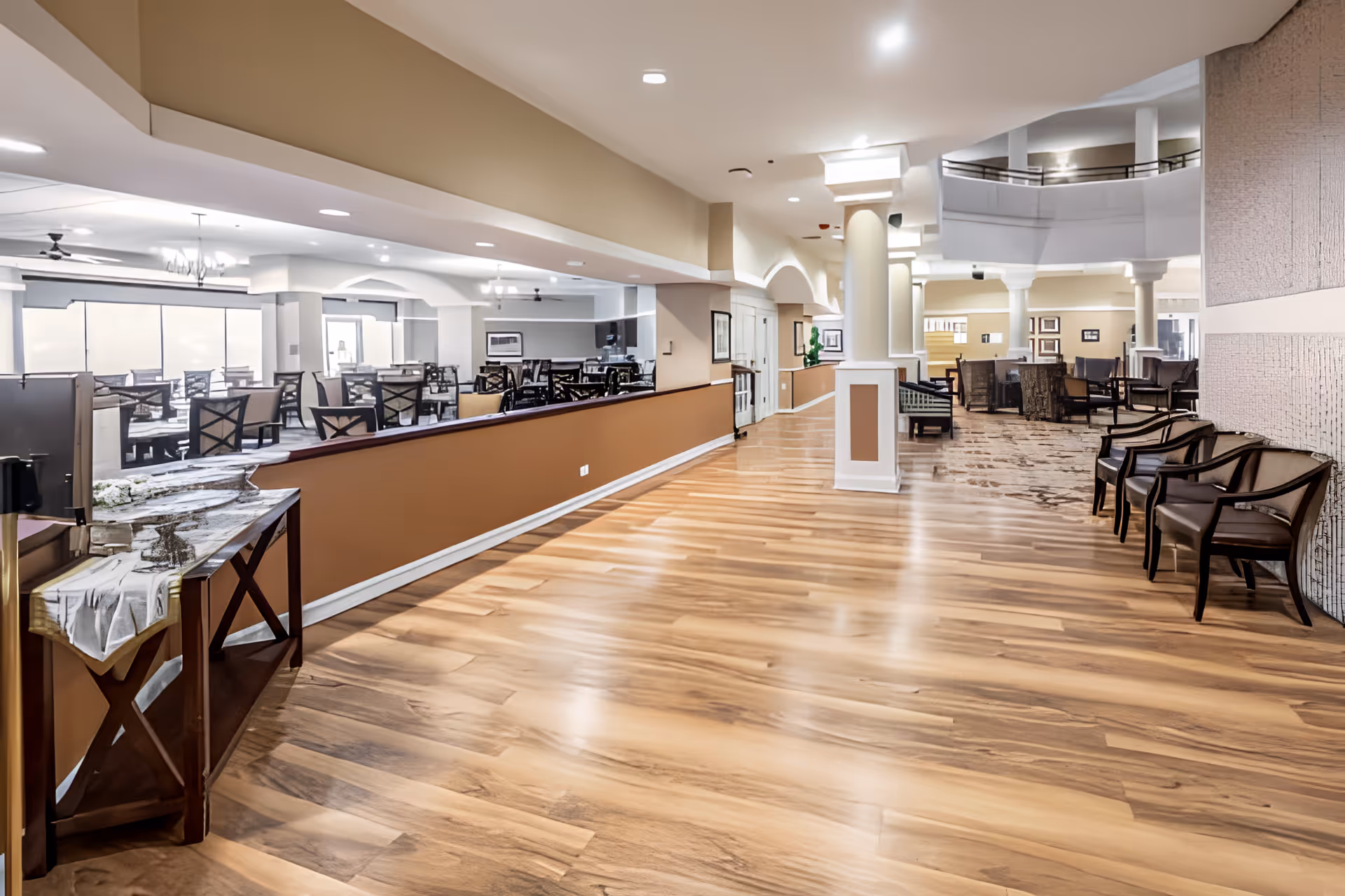 Spacious interior of a senior living facility featuring a wide hallway with polished wooden floors, columns, and seating areas with chairs. To the left, there is a dining area with multiple tables and chairs, separated by a half wall. The ceiling has recessed lighting and ceiling fans, and the overall decor is warm and inviting with neutral tones.