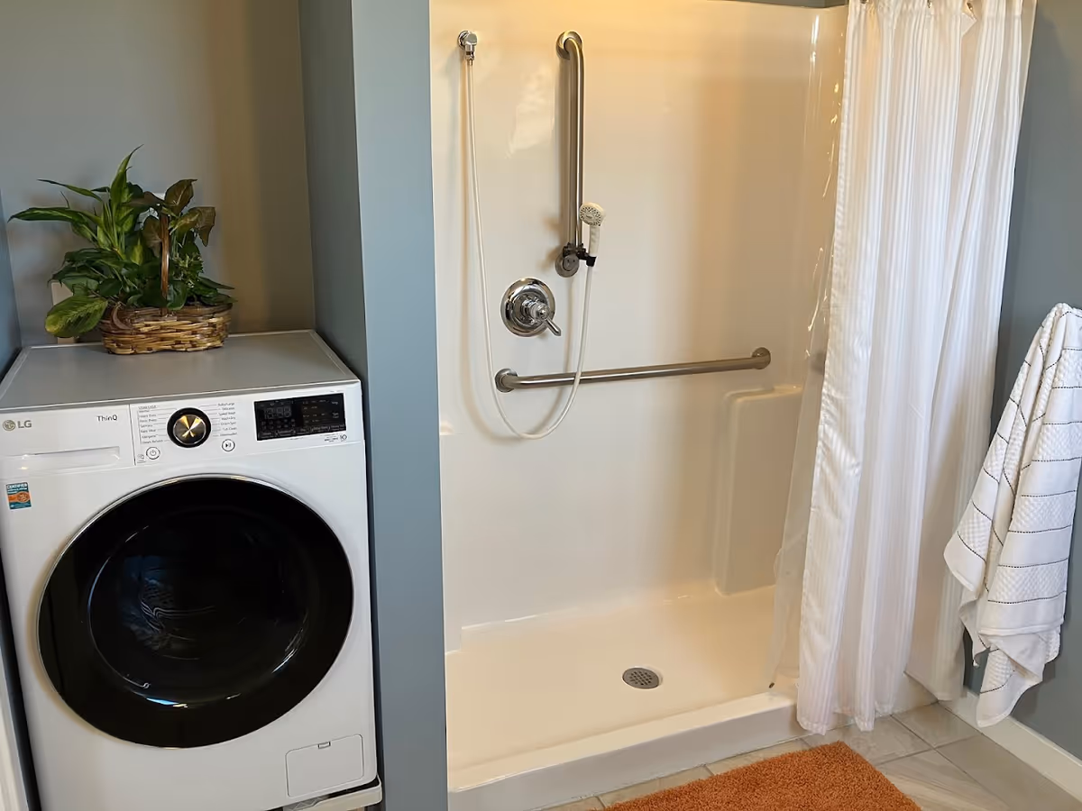 A bathroom area featuring a white walk-in shower with a handheld showerhead and grab bars. Next to the shower is a white front-loading washing machine with a green potted plant on top. A white towel hangs on a hook on the wall, and an orange bath mat is on the floor.