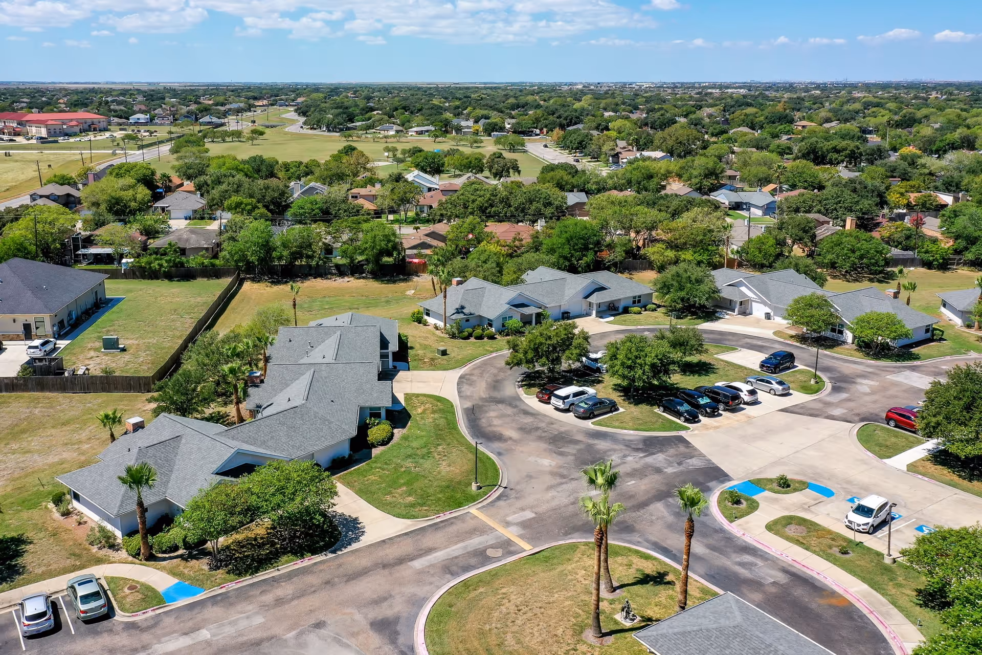Aerial view of Solstice Senior Living at Corpus Christi showing multiple single-story buildings with gray roofs arranged around a circular driveway with parked cars. The surrounding area includes green lawns, trees, and a residential neighborhood in the background under a partly cloudy sky.