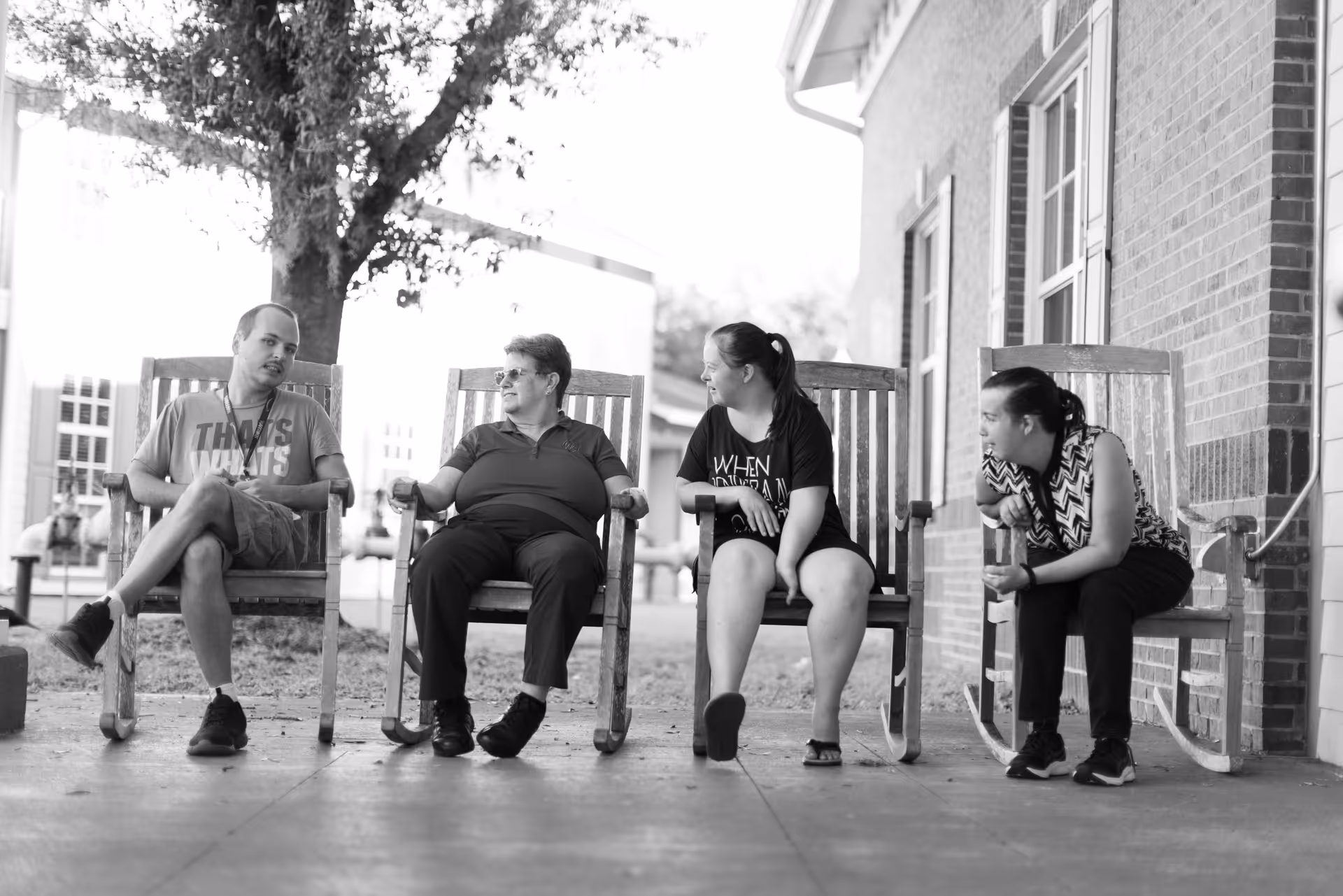Black and white photo of four people sitting on wooden rocking chairs on a porch outside a brick building, engaged in conversation and smiling.