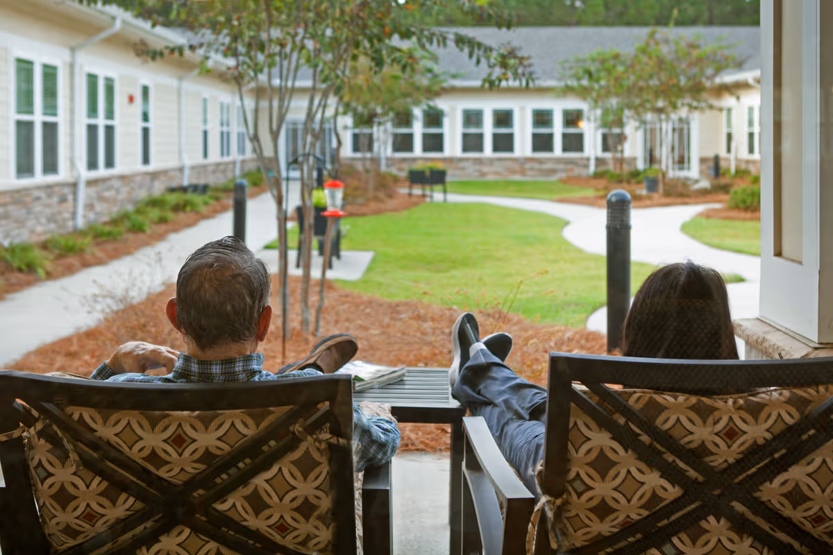 Two people sitting on cushioned outdoor chairs on a covered porch, overlooking a landscaped courtyard with a winding concrete path, small trees, and a building with many windows in the background.