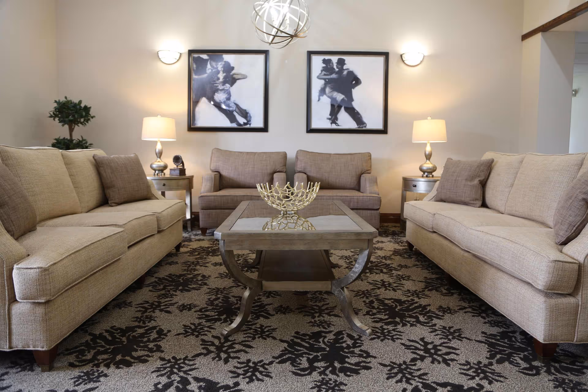 A cozy living room with two beige sofas facing each other and two matching armchairs against the back wall. A glass-top coffee table with a decorative gold bowl is centered on a patterned carpet. Two framed black and white pictures of dancing couples hang on the wall above the armchairs. Two table lamps on side tables and wall sconces provide warm lighting.