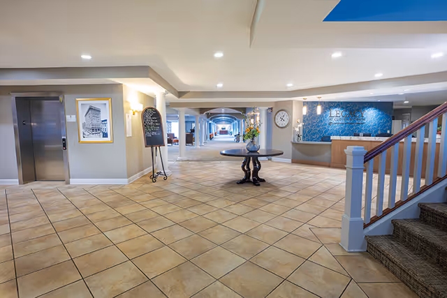 Spacious and well-lit interior lobby area of Legacy at Cranberry Landing featuring tiled floors, a round table with a flower vase in the center, an elevator on the left, a reception desk on the right with a blue patterned wall behind it, and a staircase with carpeted steps on the right side.