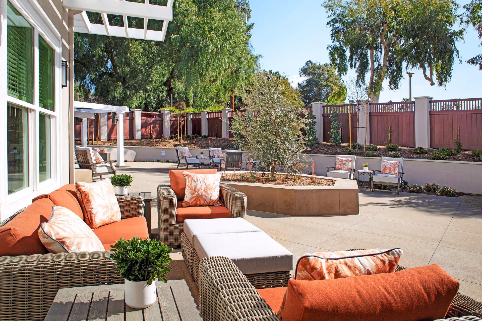 Outdoor patio area with wicker furniture including sofas and chairs with orange cushions and patterned pillows, small tables with potted plants, a central planter with a tree, surrounded by a wooden fence and tall trees in the background under a clear blue sky.