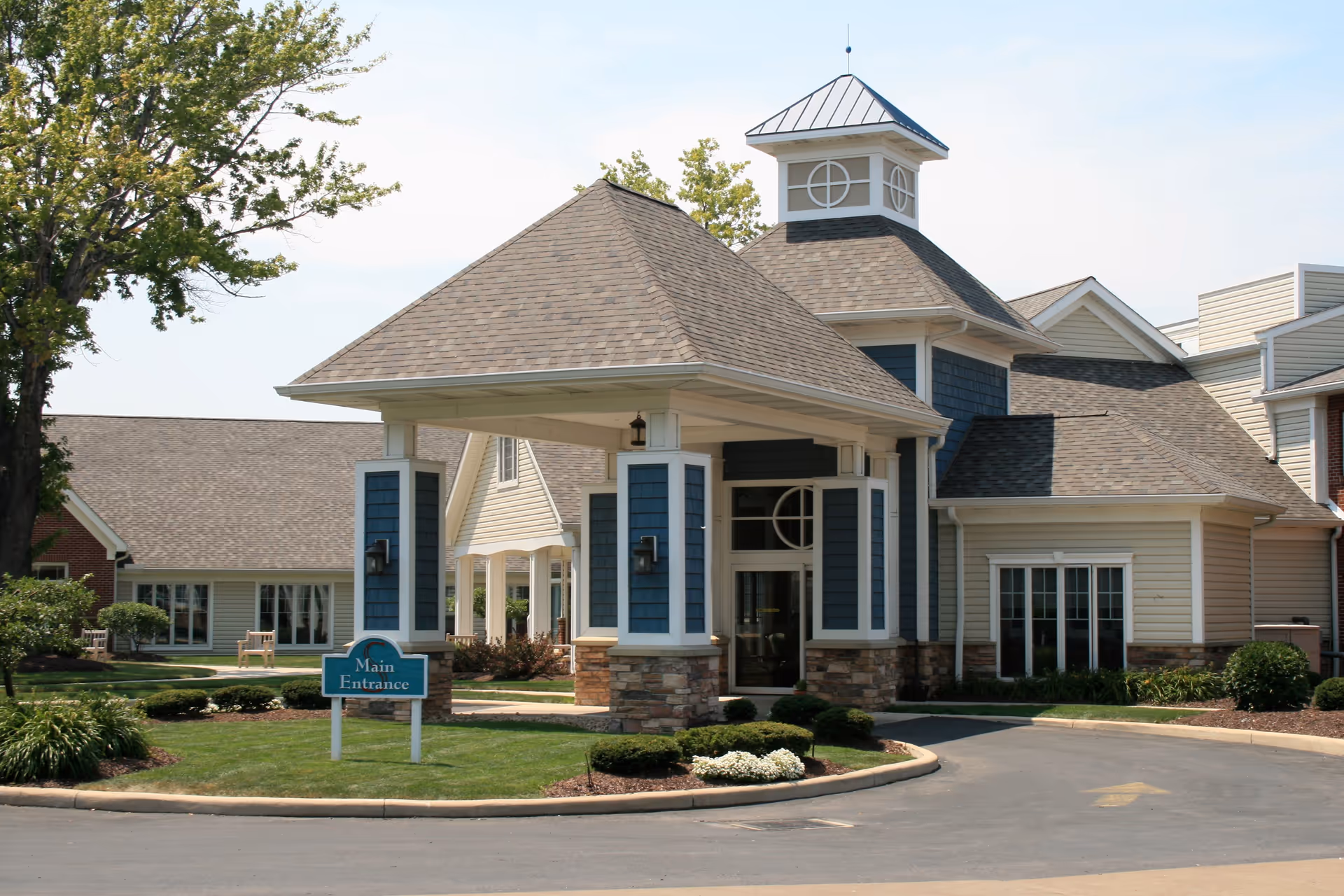 Exterior view of the main entrance to Sprenger Health Care Anchor Lodge Retirement Village, featuring a covered driveway with blue and beige siding, stone accents, and a small landscaped area with bushes and flowers.